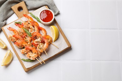 Photo of Delicious grilled shrimp skewers served with sauce and lemon on white tiled table, top view. Space for text