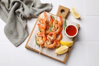 Photo of Delicious grilled shrimp skewers served with sauce and lemon on white tiled table, top view