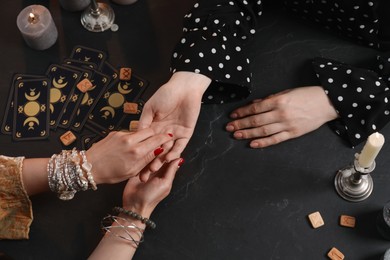 Fortune teller reading lines on woman's palm at black table, top view. Chiromancy