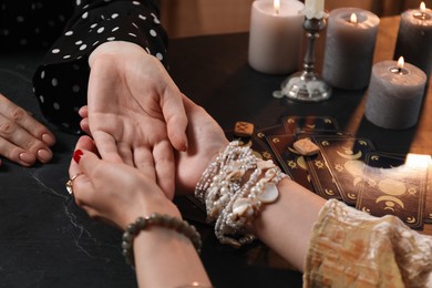 Fortune teller reading lines on woman's palm at black table, closeup. Chiromancy