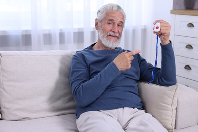 Photo of Happy senior man pointing at emergency call button on sofa indoors