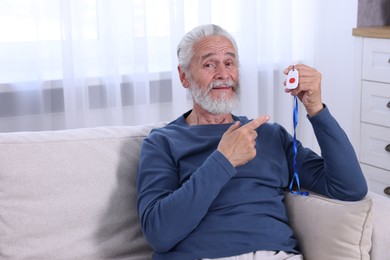 Photo of Happy senior man pointing at emergency call button on sofa indoors