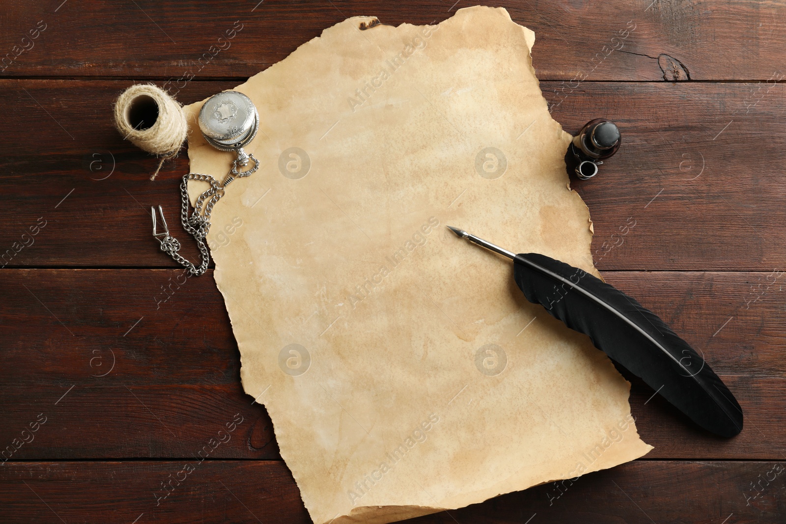 Photo of Sheet of old parchment paper, black feather, inkwell, rope and pocket chain clock on wooden table, flat lay