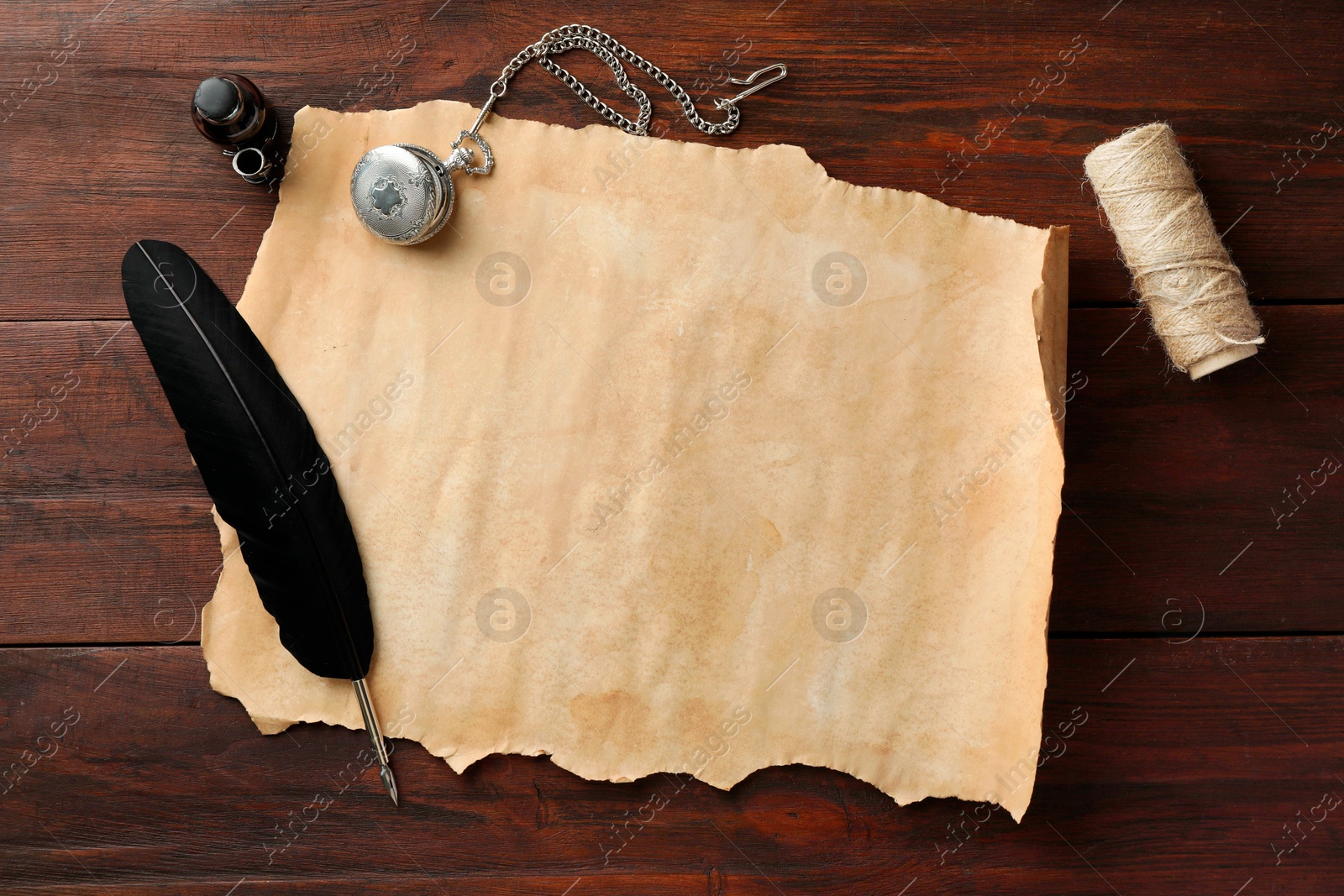 Photo of Sheet of old parchment paper, black feather, inkwell, rope and pocket chain clock on wooden table, flat lay