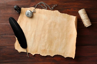 Sheet of old parchment paper, black feather, inkwell, rope and pocket chain clock on wooden table, flat lay