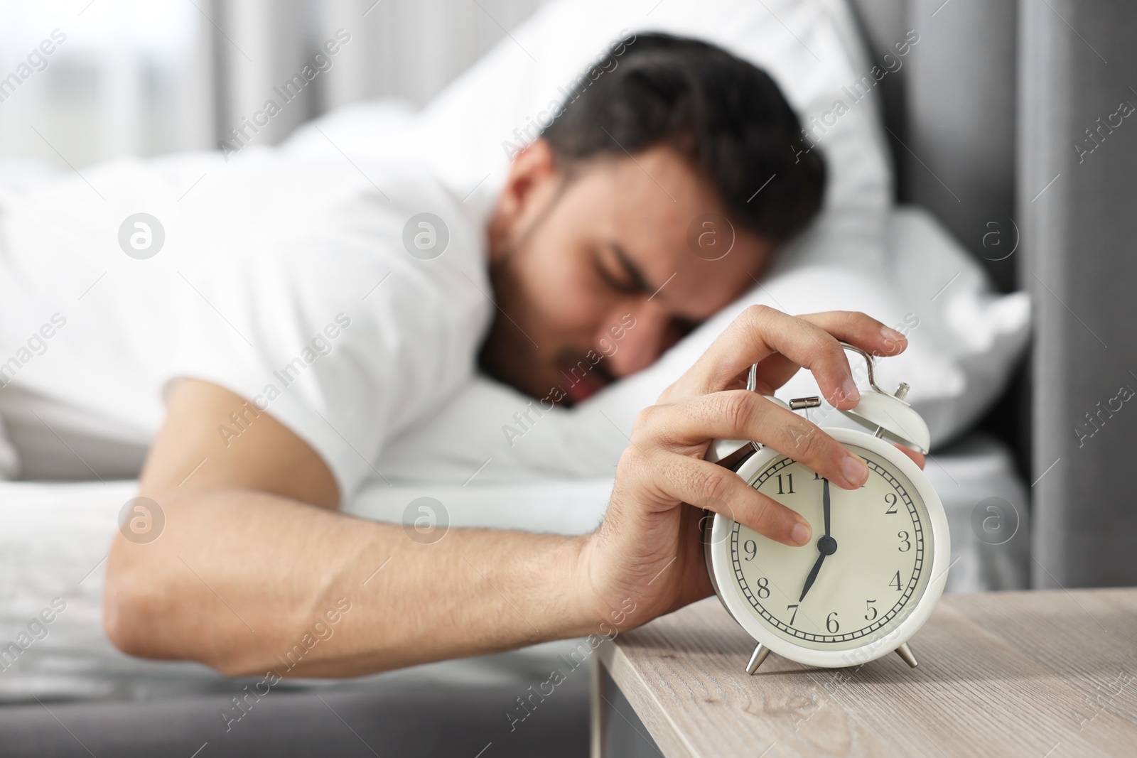 Photo of Sleepy man turning off alarm clock in bedroom at morning, selective focus