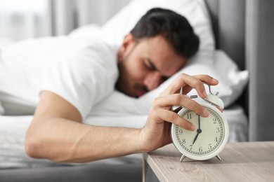 Photo of Sleepy man turning off alarm clock in bedroom at morning, selective focus