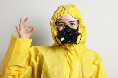 Photo of Worker in respirator, protective suit and glasses showing OK gesture on grey background