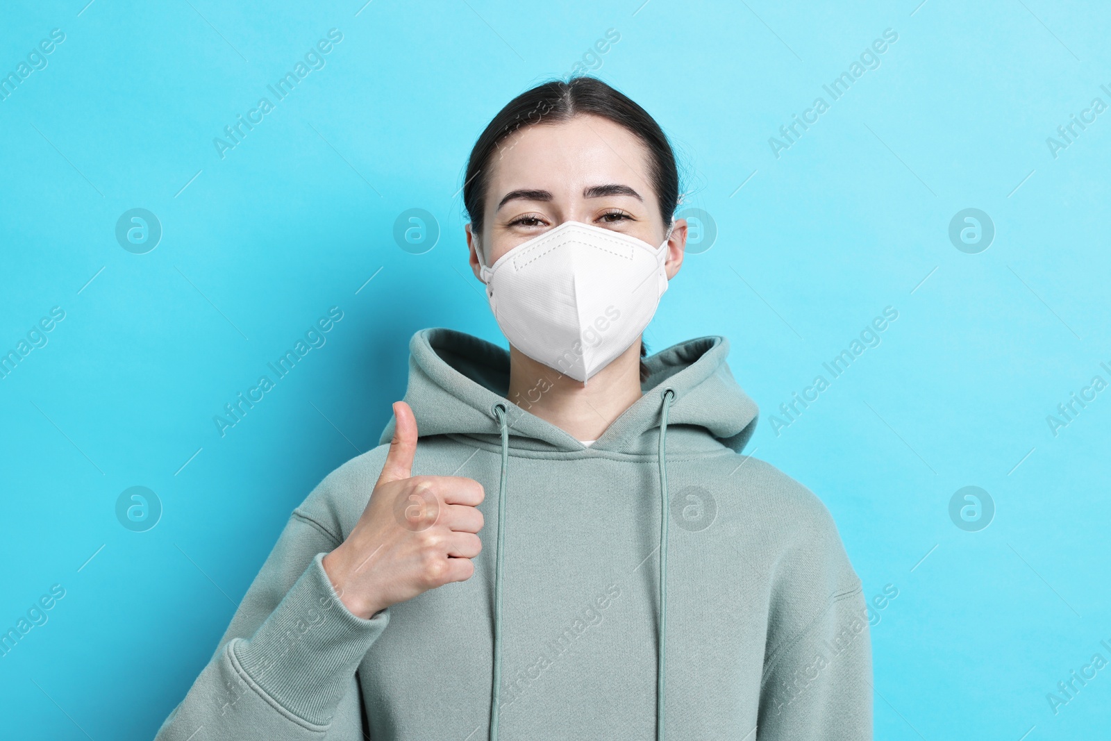 Photo of Woman in respirator mask showing thumbs up on light blue background