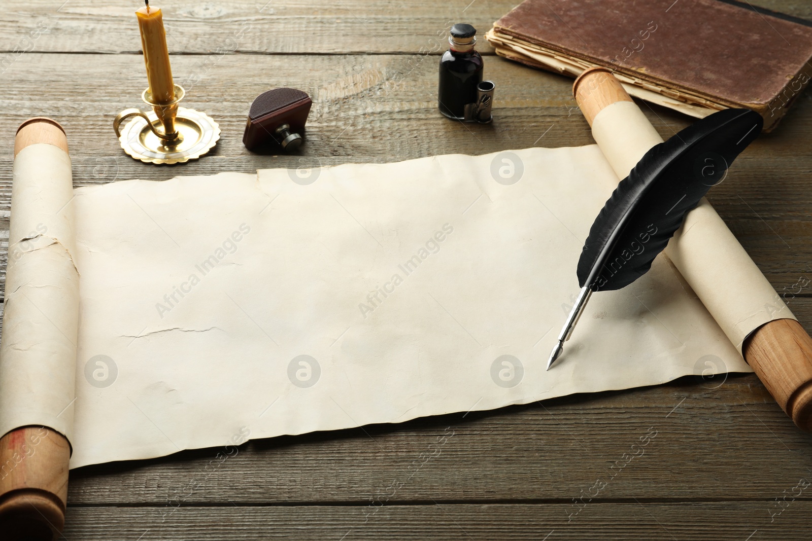 Photo of Sheet of old parchment paper, wax stamp, black feather, inkwell, candle and vintage book on wooden table