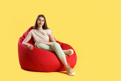Photo of Beautiful young woman sitting on red bean bag chair against yellow background, space for text
