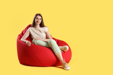 Beautiful young woman sitting on red bean bag chair against yellow background, space for text