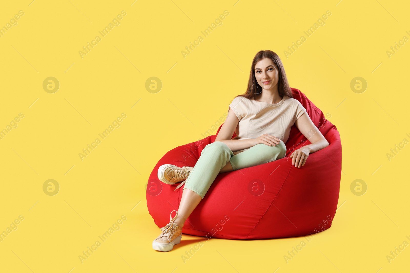 Photo of Beautiful young woman sitting on red bean bag chair against yellow background, space for text