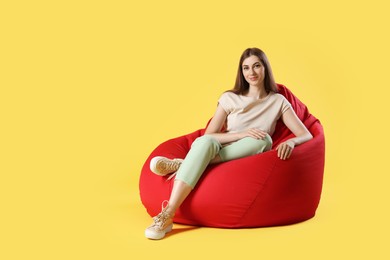 Photo of Beautiful young woman sitting on red bean bag chair against yellow background, space for text