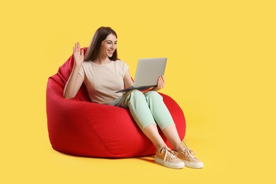 Photo of Smiling woman with laptop having online meeting on red bean bag chair against yellow background