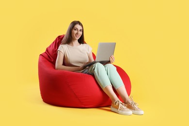 Photo of Smiling woman with laptop sitting on red bean bag chair against yellow background