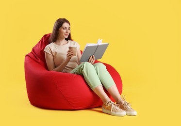 Photo of Smiling woman with book and paper cup of drink sitting on red bean bag chair against yellow background