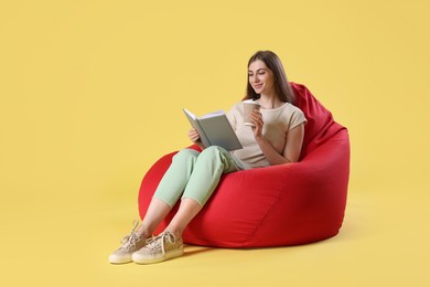 Photo of Smiling woman with book and paper cup of drink sitting on red bean bag chair against yellow background