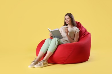 Smiling woman with book and paper cup of drink sitting on red bean bag chair against yellow background