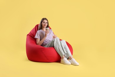 Beautiful young woman with paper cup of drink sitting on red bean bag chair against yellow background, space for text