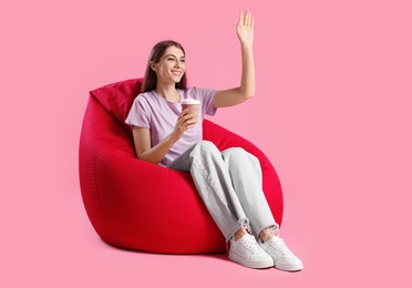 Photo of Smiling woman with paper cup of drink sitting on red bean bag chair against pink background