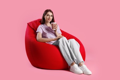 Beautiful young woman with paper cup of drink sitting on red bean bag chair against pink background