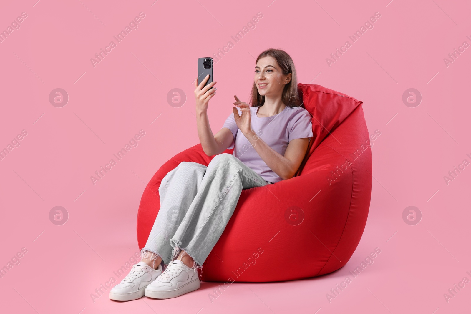 Photo of Smiling woman with smartphone having online meeting on red bean bag chair against pink background