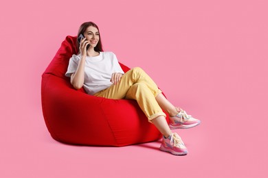 Photo of Smiling woman talking on smartphone while sitting on red bean bag chair against pink background