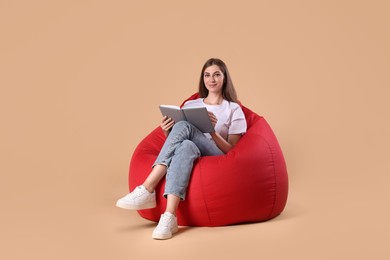 Photo of Beautiful young woman reading book on red bean bag chair against beige background