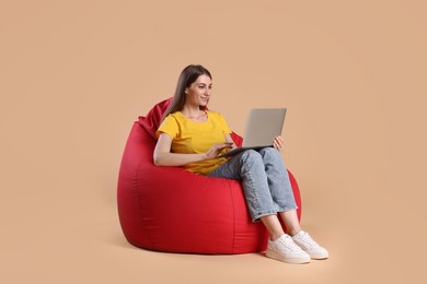 Photo of Smiling woman using laptop on red bean bag chair against beige background