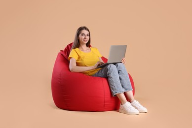 Photo of Beautiful young woman using laptop on red bean bag chair against beige background