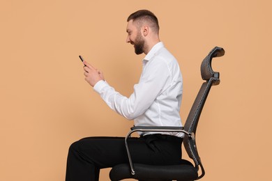 Photo of Man with good posture sitting on chair and using smartphone against pale orange background