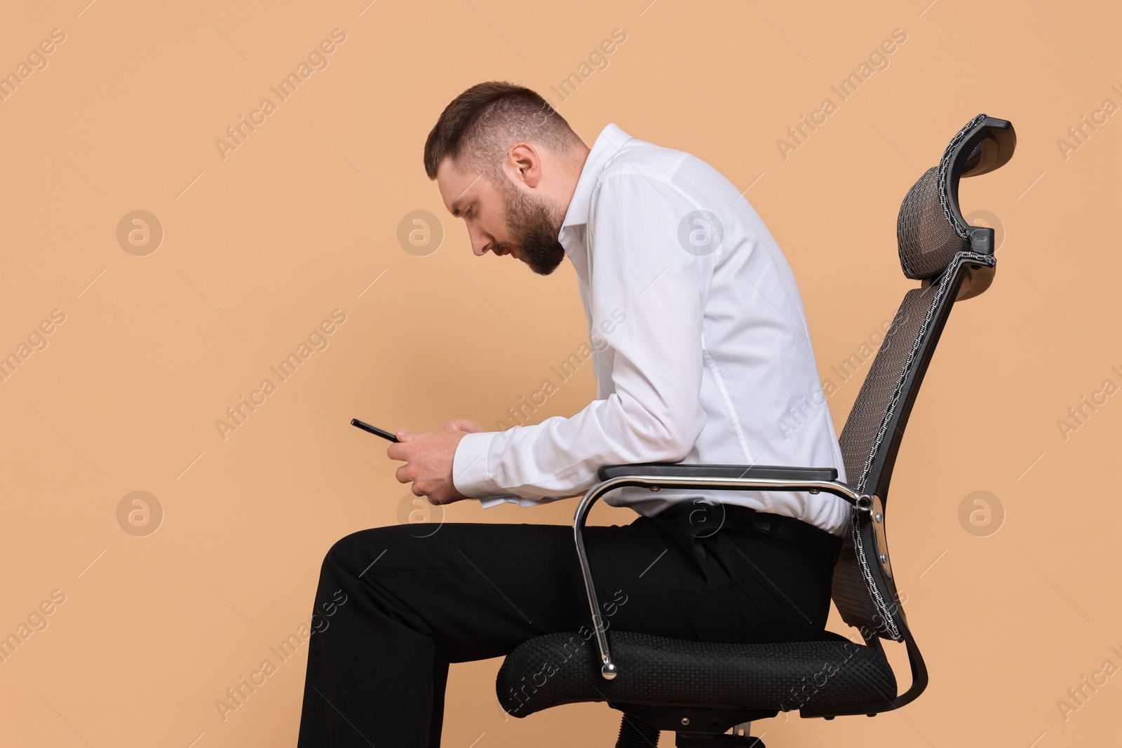 Photo of Man with poor posture sitting on chair and using smartphone against pale orange background