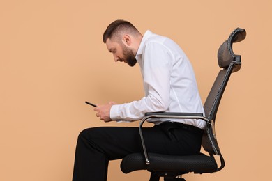 Photo of Man with poor posture sitting on chair and using smartphone against pale orange background