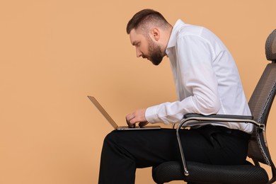 Man with poor posture sitting on chair and using laptop against pale orange background, space for text