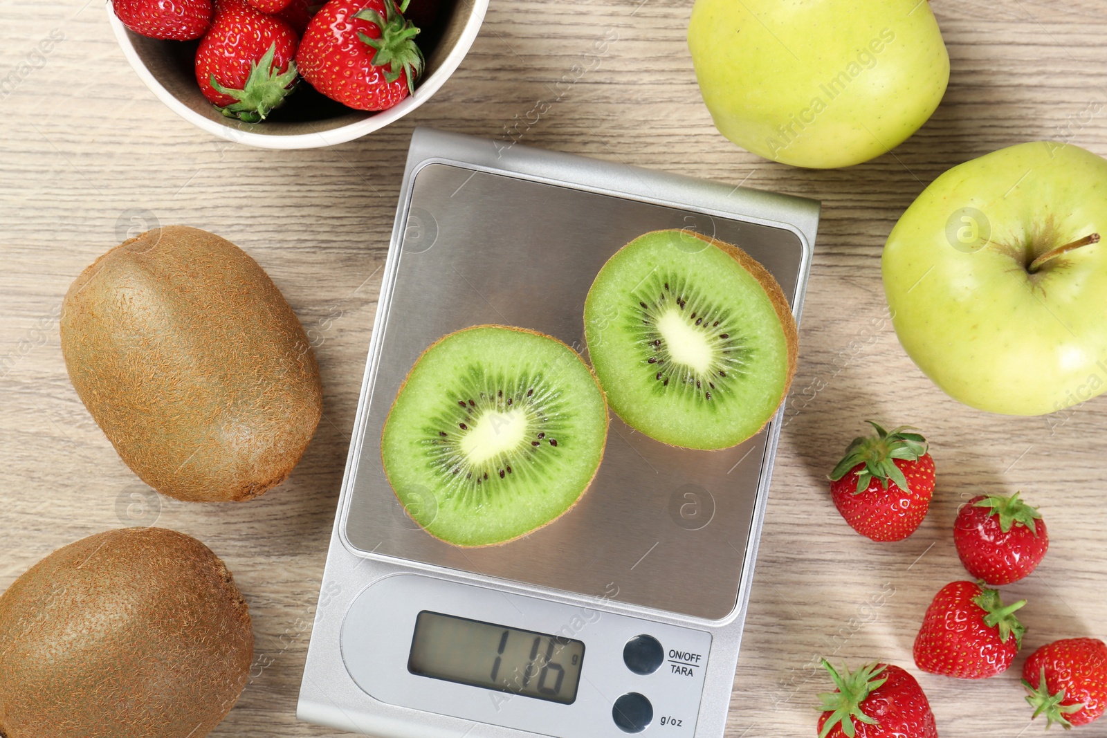 Photo of Kitchen scale with halves of kiwi among apples and strawberries on light wooden table, flat lay