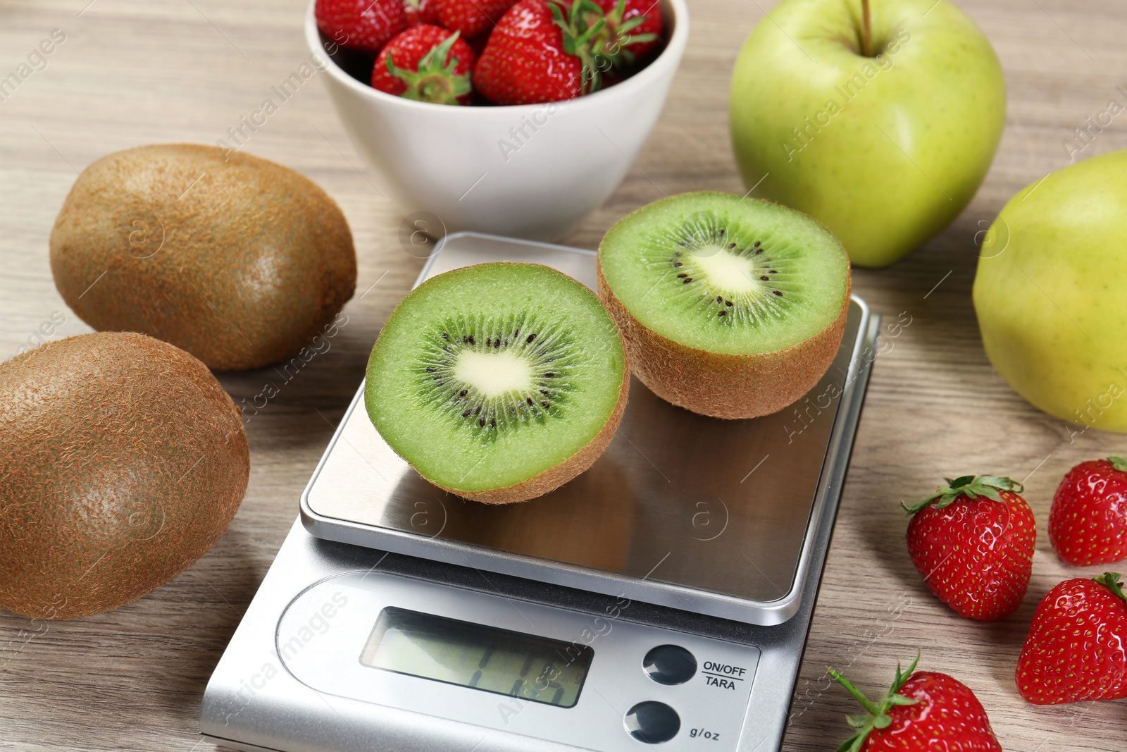 Photo of Kitchen scale with halves of kiwi among apples and strawberries on light wooden table, closeup