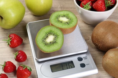 Photo of Kitchen scale with halves of kiwi among apples and strawberries on light wooden table, closeup