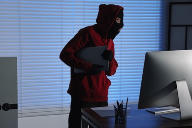 Photo of Thief wearing balaclava stealing laptop in office at night. Burglary