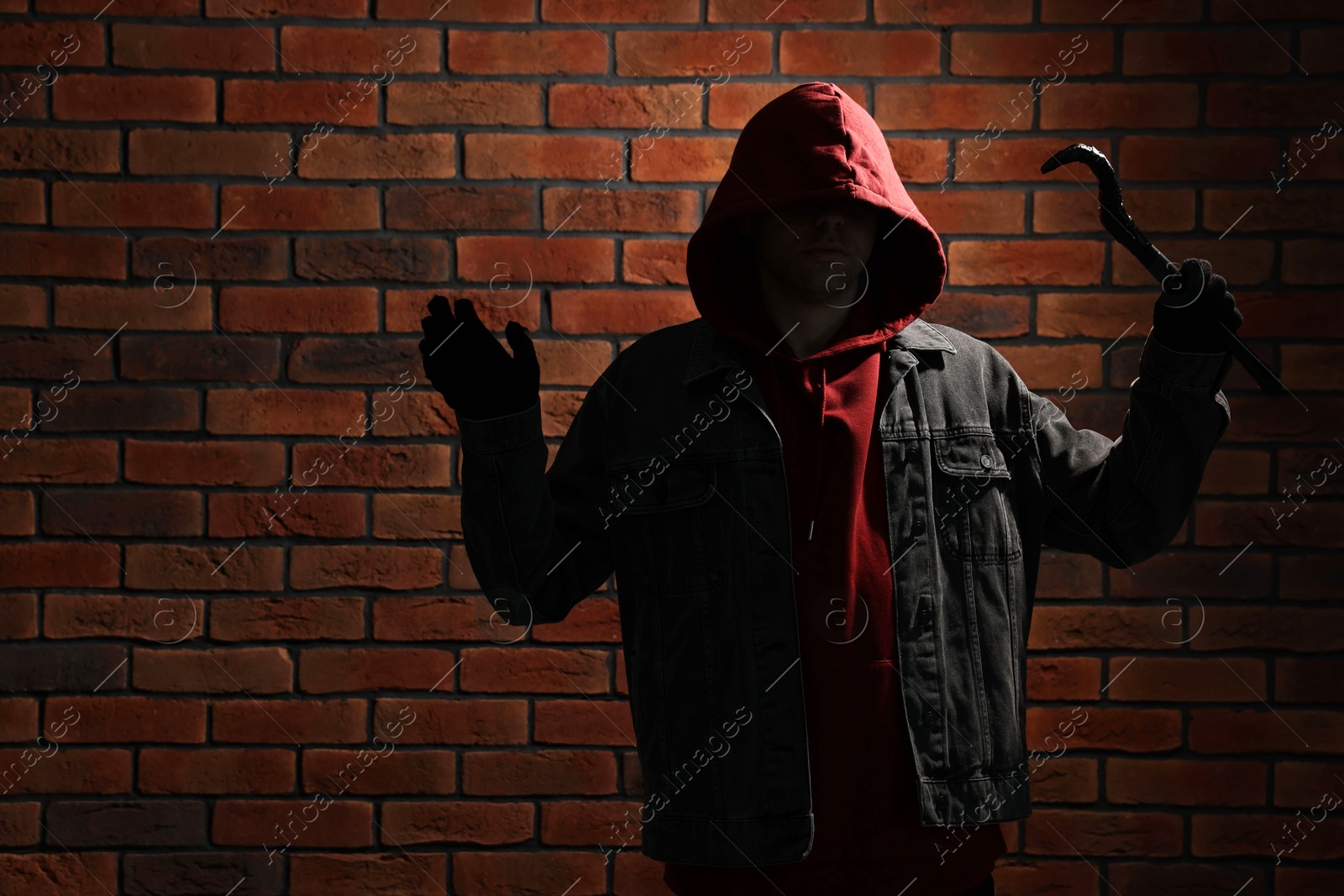 Photo of Thief in hoodie raising hands with crowbar against red brick wall. Space for text