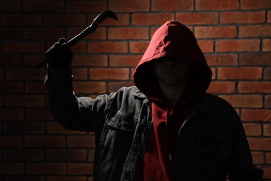 Thief in hoodie raising hand with crowbar against red brick wall