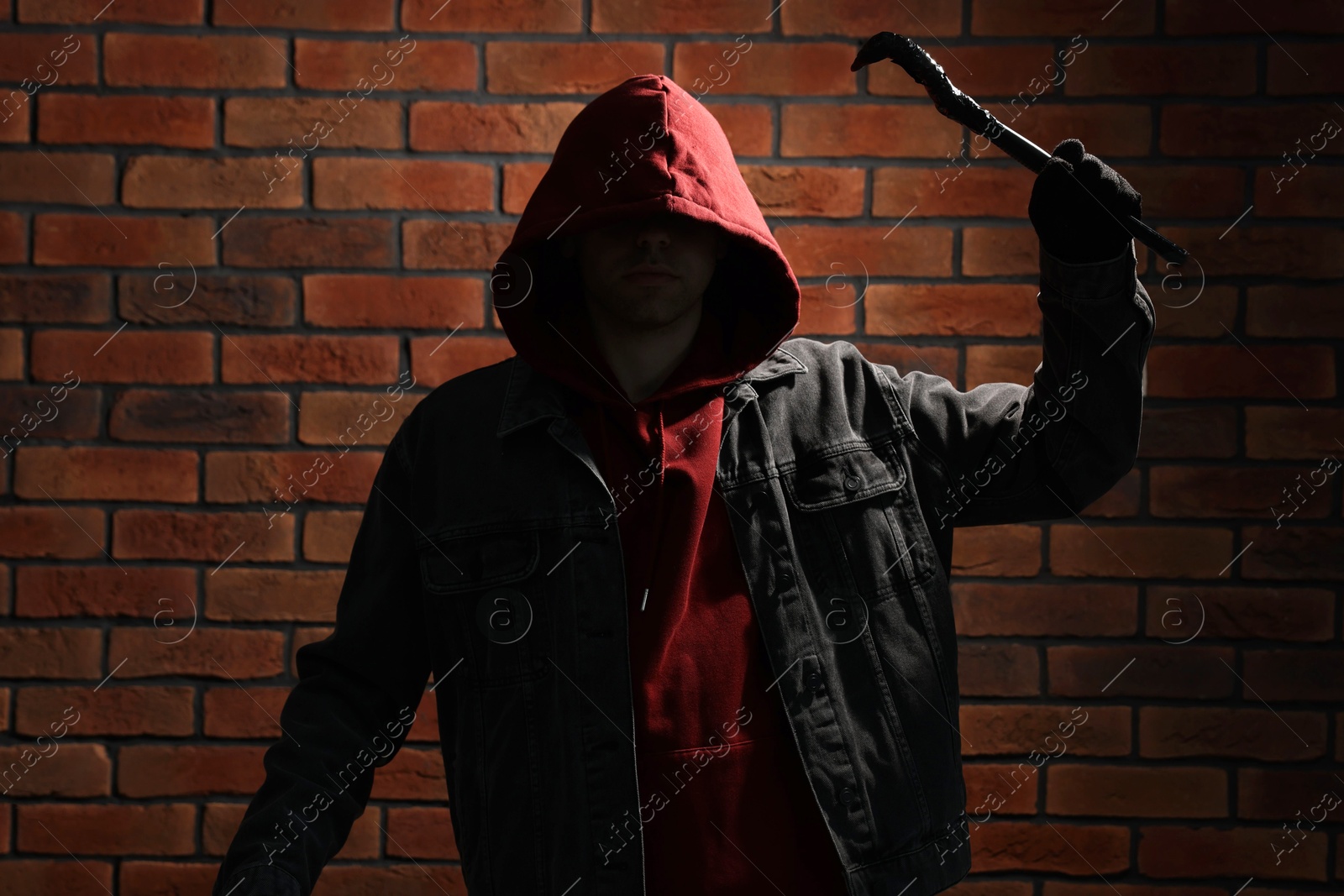 Photo of Thief in hoodie raising hand with crowbar against red brick wall