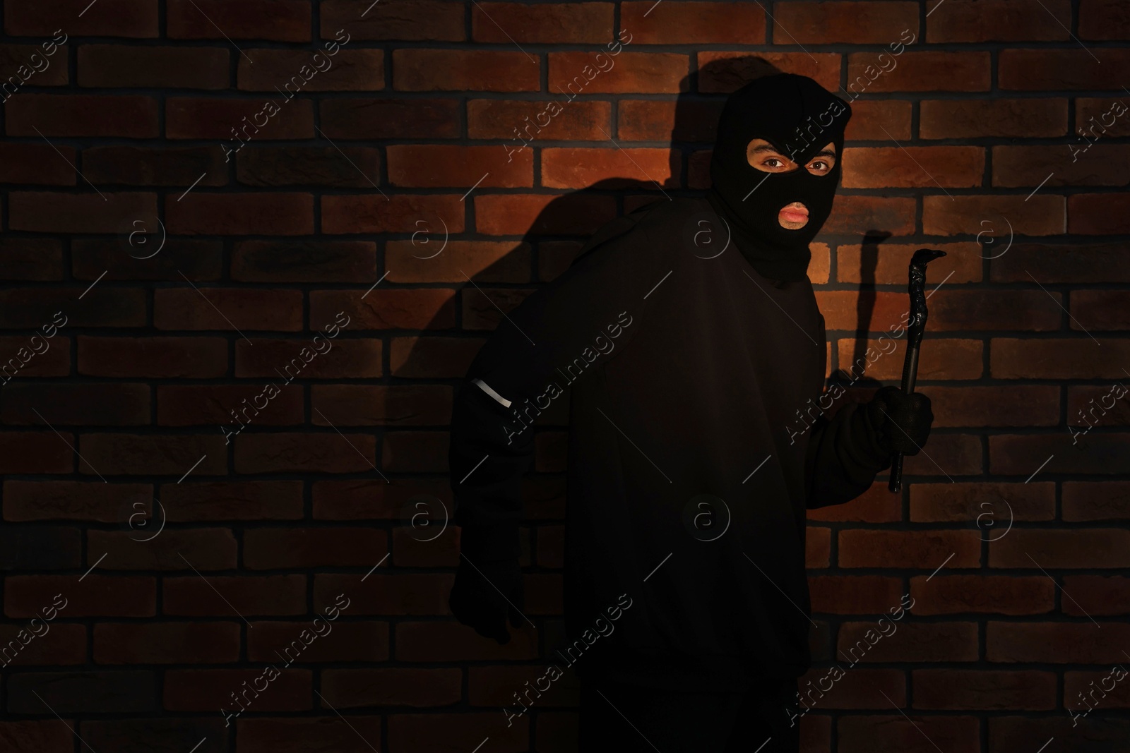 Photo of Thief in balaclava with crowbar against red brick wall. Space for text