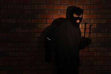 Thief in balaclava with crowbar against red brick wall. Space for text