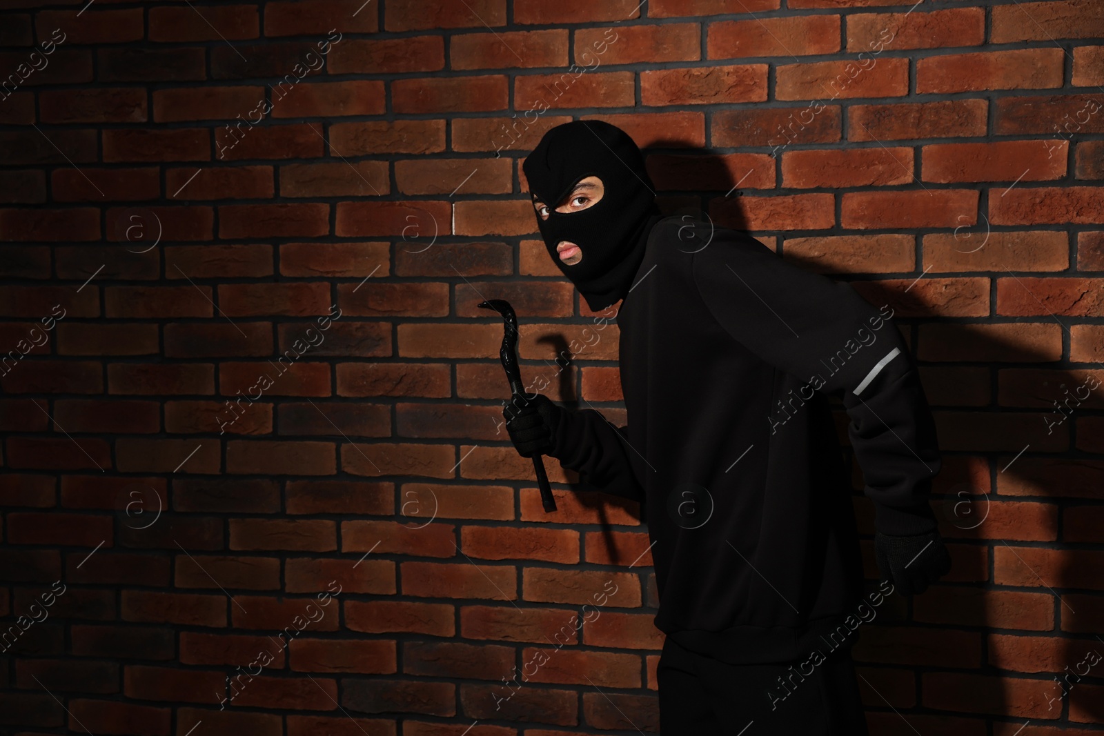 Photo of Thief in balaclava with crowbar against red brick wall. Space for text