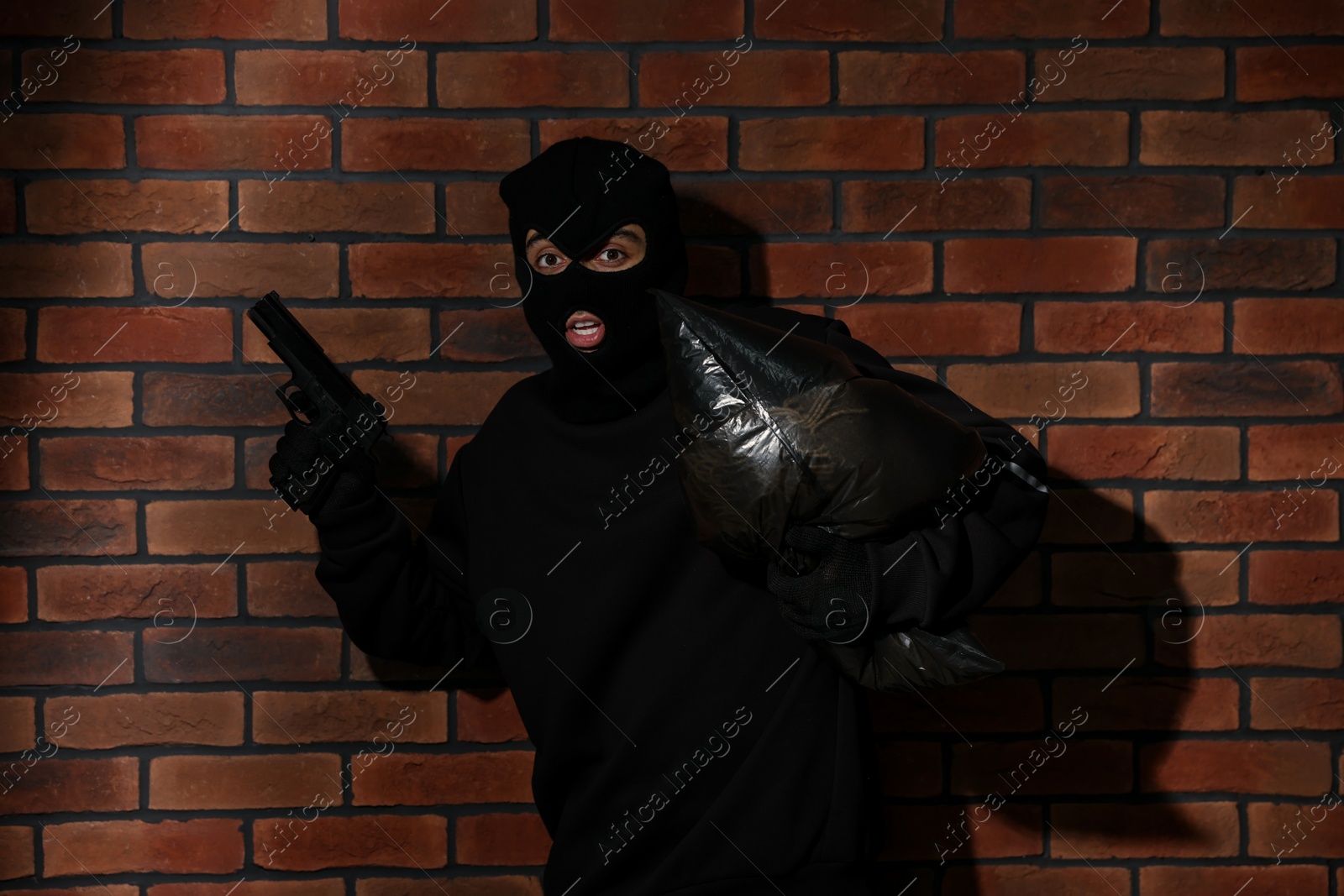 Photo of Emotional thief in balaclava with gun and bag against red brick wall