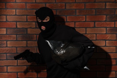 Photo of Thief in balaclava with gun and bag against red brick wall