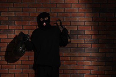 Photo of Emotional thief in balaclava raising hands with crowbar and bag against red brick wall. Space for text