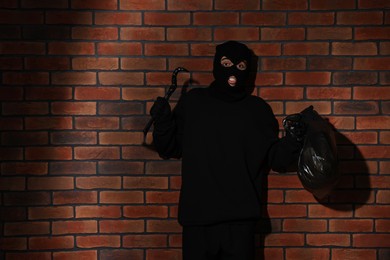 Photo of Emotional thief in balaclava raising hands with crowbar and bag against red brick wall. Space for text