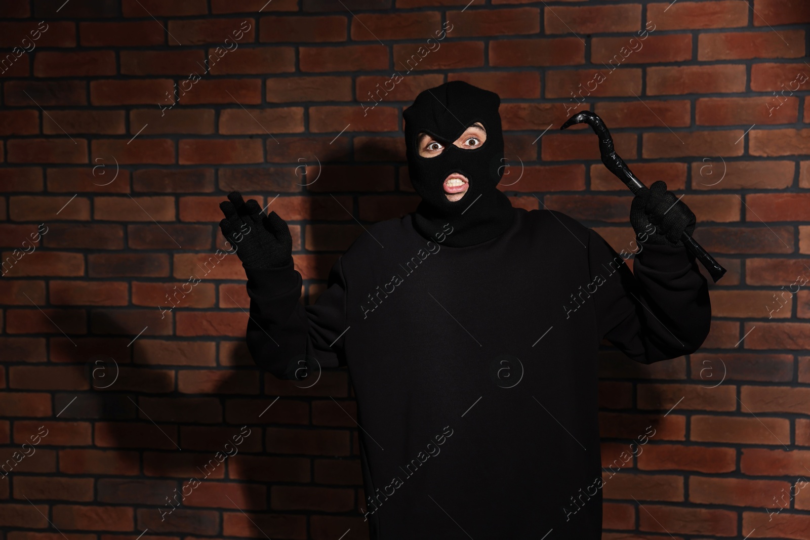 Photo of Emotional thief in balaclava raising hands with crowbar against red brick wall